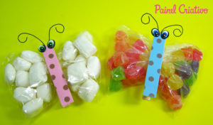 Duas embalagens de balas em formato de borboleta feitas com saquinhos plásticos transparentes preenchidos com doces coloridos, decoradas com tiras de EVA rosa e azul representando o corpo da borboleta, olhos grandes e antenas desenhadas em fundo amarelo vivo.