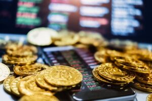 Várias moedas douradas de Bitcoin sobre um celular com gráfico de criptomoedas ao fundo.